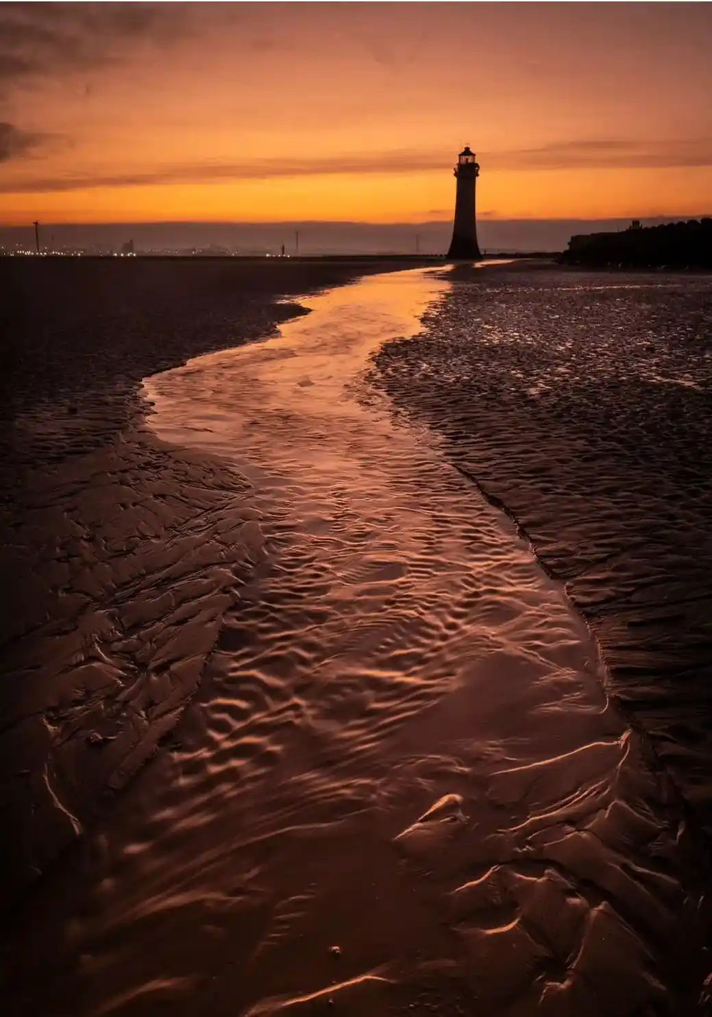 A serene sunset photograph shows a lighthouse silhouetted against an orange and yellow sky, with a narrow channel of water…