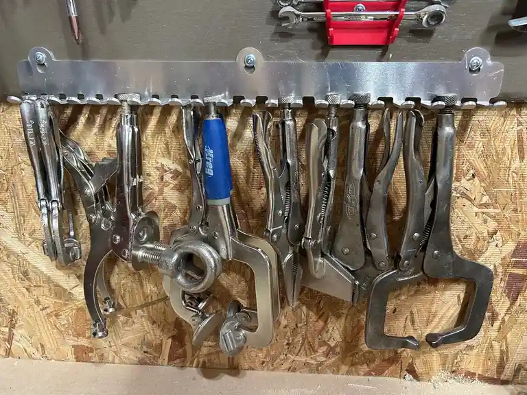 A metal rack mounted on a particleboard wall holds a variety of metal clamps and pliers, mostly in silver or gray tones…