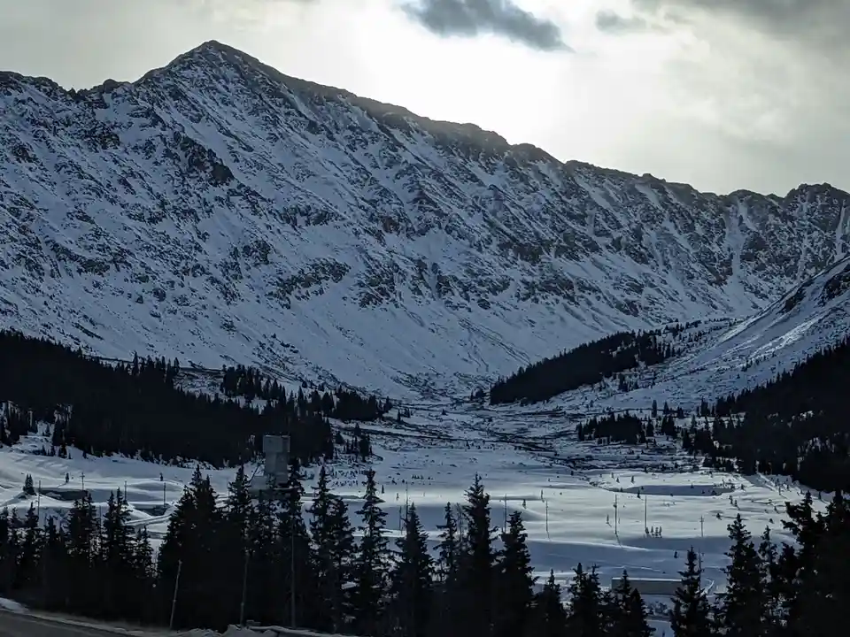 A wide shot of a snow-covered mountain valley with ski slopes, evergreen trees, and a cloudy sky. The overlaid text reads…