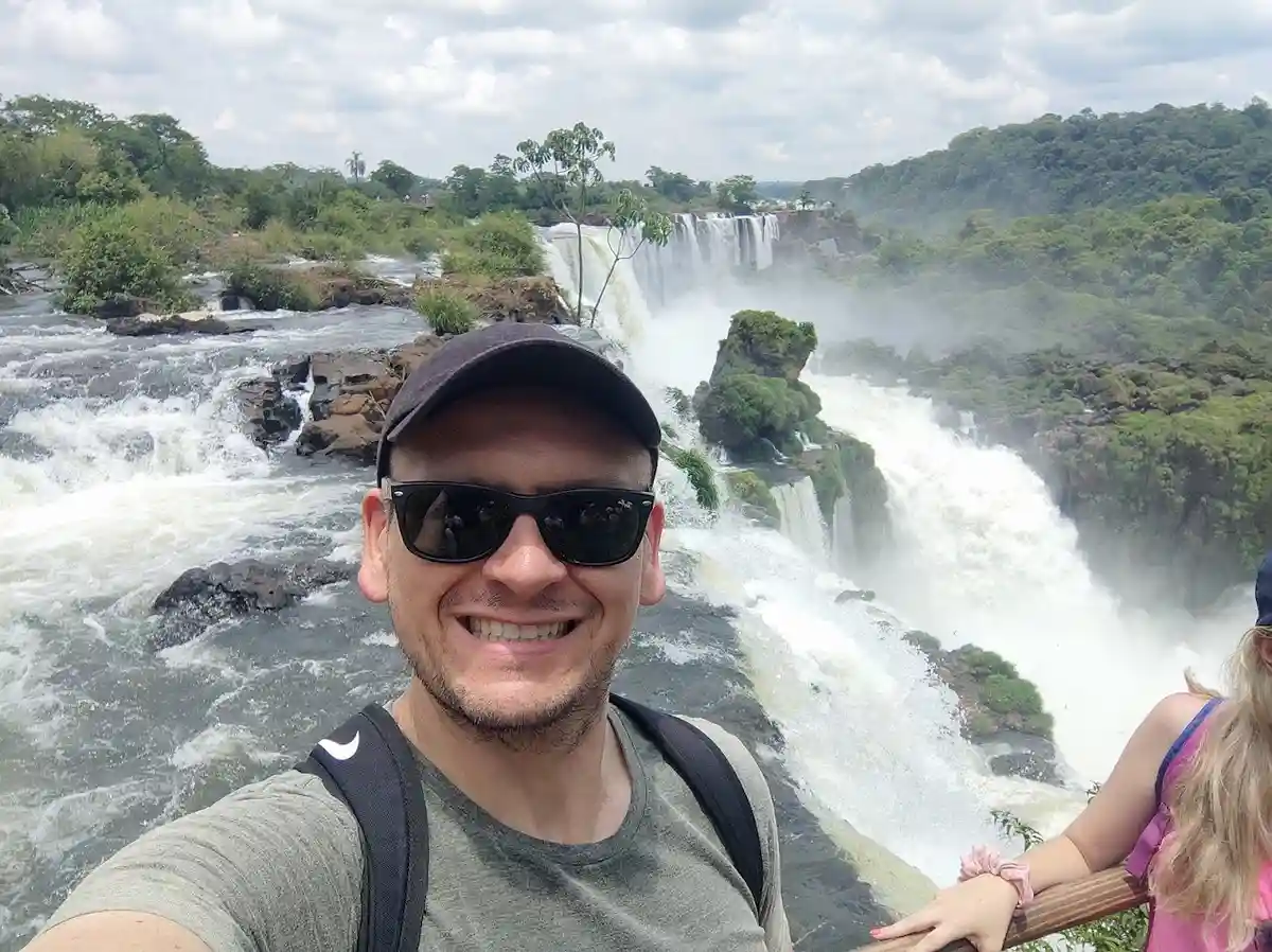 A man wearing a black cap and sunglasses smiles broadly at the camera while taking a selfie in front of a massive, misty…