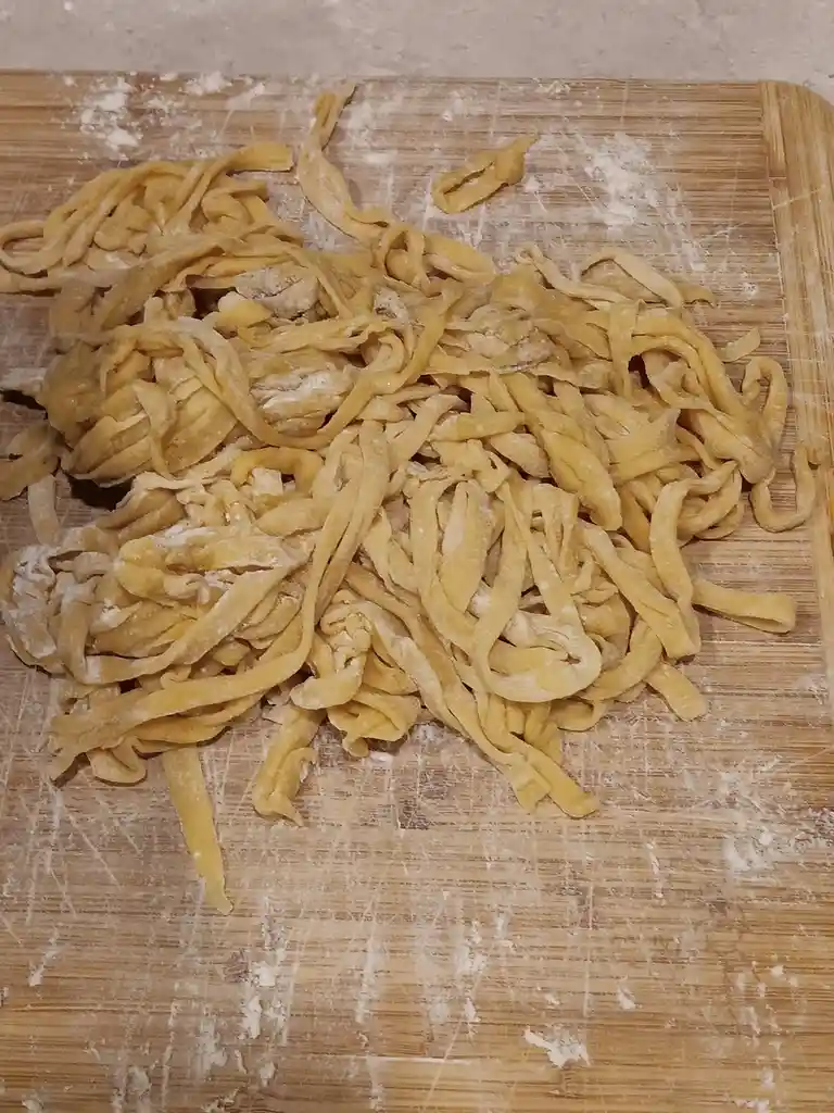 A pile of homemade, uncooked pasta noodles lies scattered on a well-used wooden cutting board dusted with flour. The pasta…