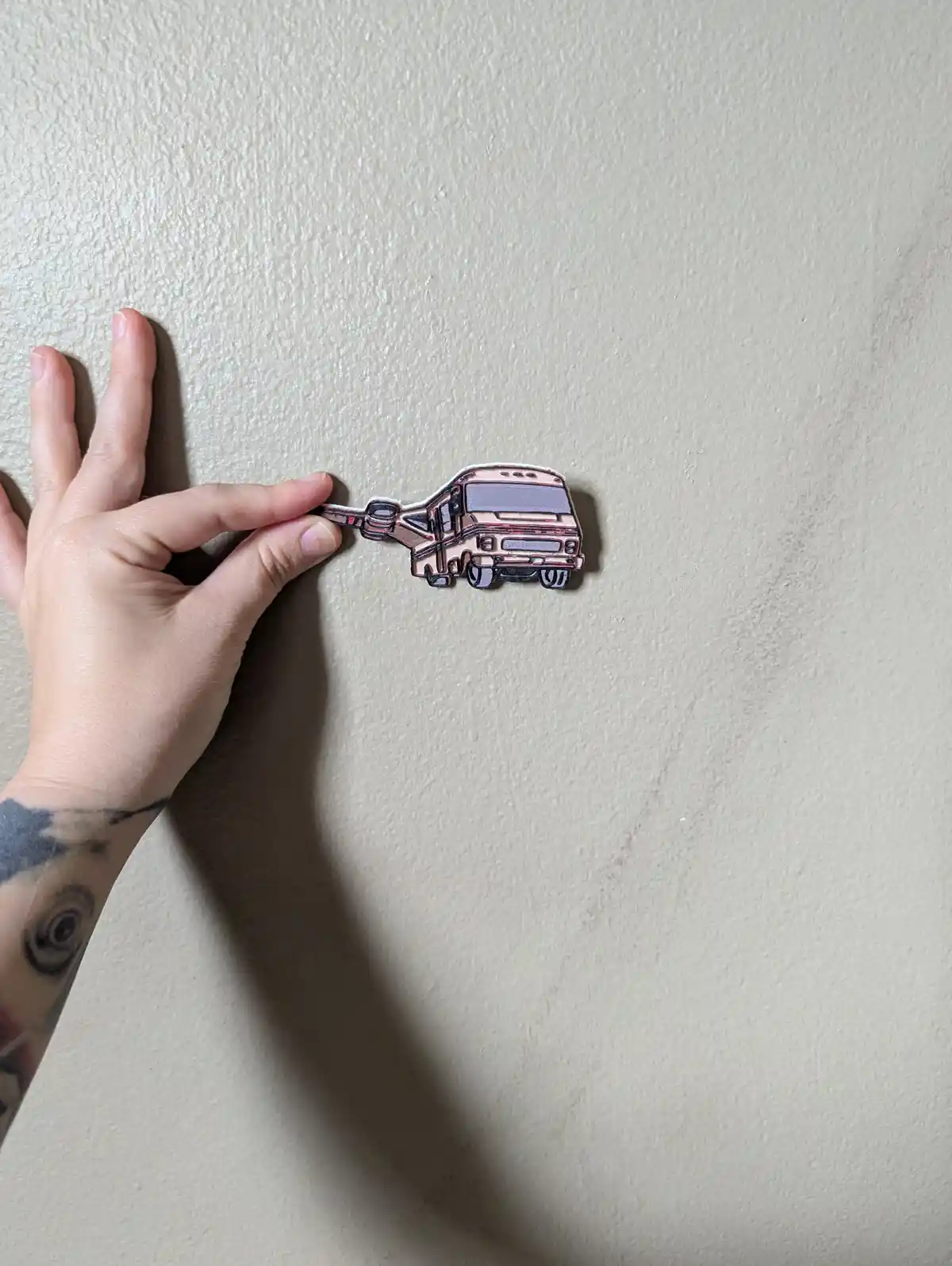 A hand with tattoos holds a small, metallic-looking RV-shaped magnet against a textured beige wall. The magnet is designed…