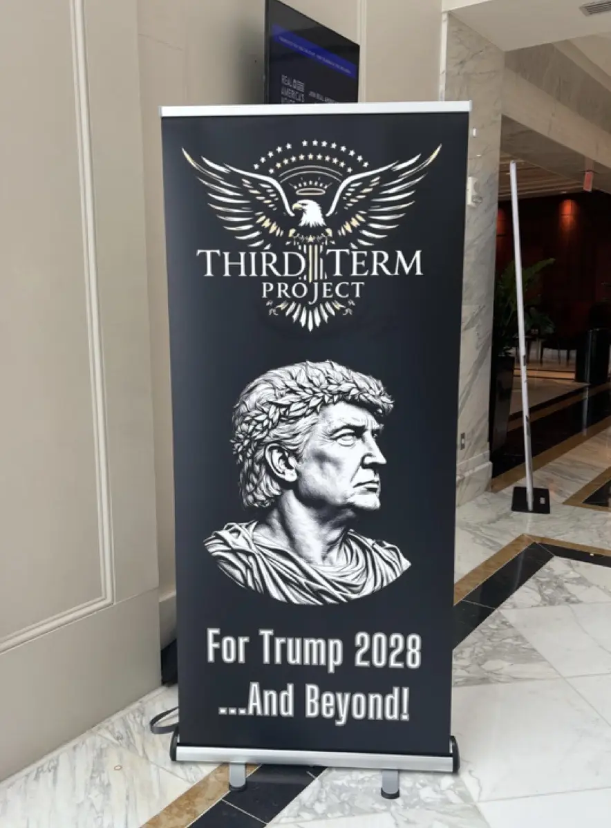 A black banner stand in a marble lobby displays a logo with an eagle and stars above the text 'THIRD TERM PROJECT'. Below, a…