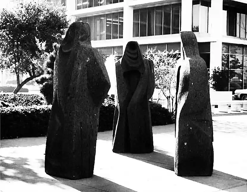 a black and white photo shows three abstract, hooded stone statues standing in a row on a paved plaza in front of a modern…