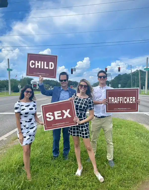 Four people stand in a grassy area beside a road holding red signs with white text. One sign reads 'CHILD', another 'SEX'…
