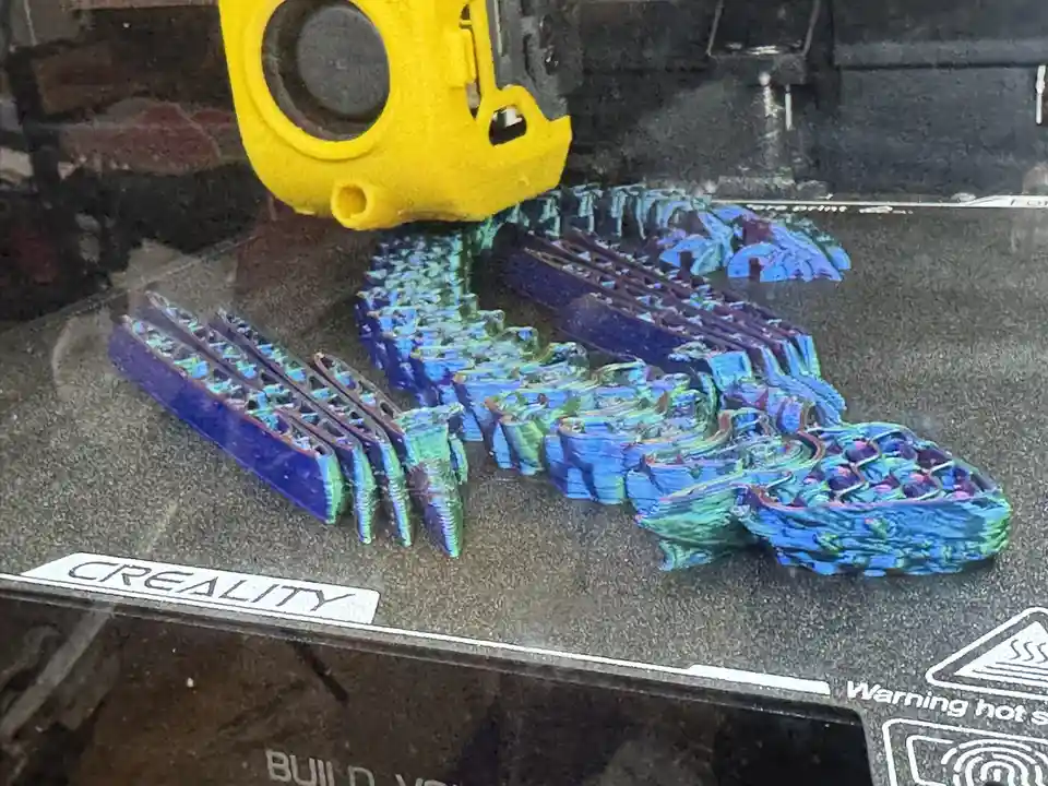 A close-up photo of a 3D printer's print bed shows a partially completed, colorful, and visibly flawed object resembling a…