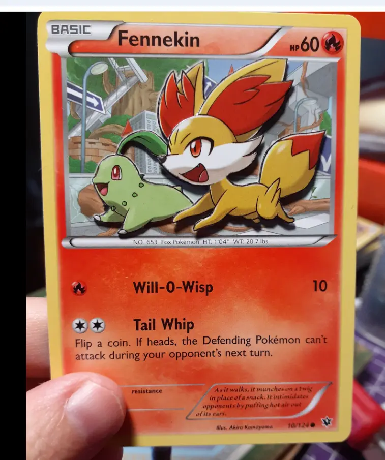 A hand holds a Pokémon trading card featuring Fennekin, a small fox-like creature with red-tipped ears and tail, alongside a…