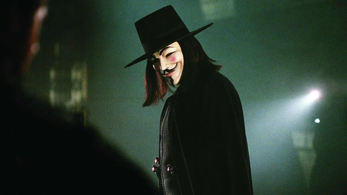 A person wearing a black top hat and a white Guy Fawkes mask with a wide grin, dressed in a black coat, stands in a dimly…