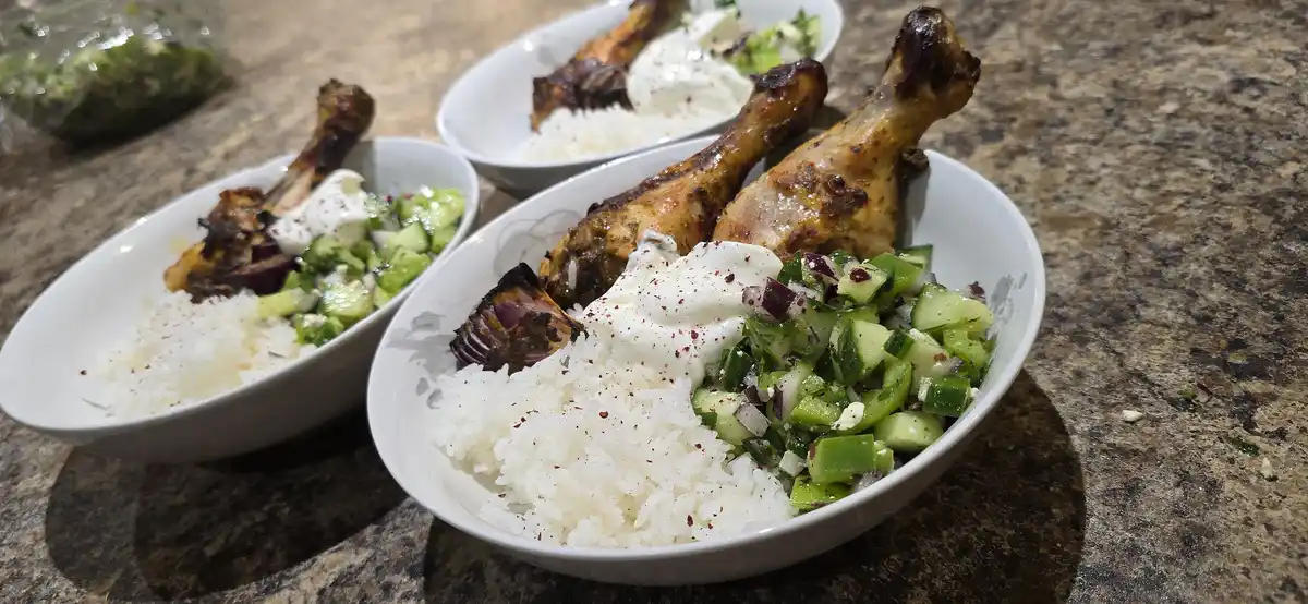 Three white bowls on a granite countertop contain grilled chicken drumsticks, white rice, and a cucumber-tomato-onion salad…