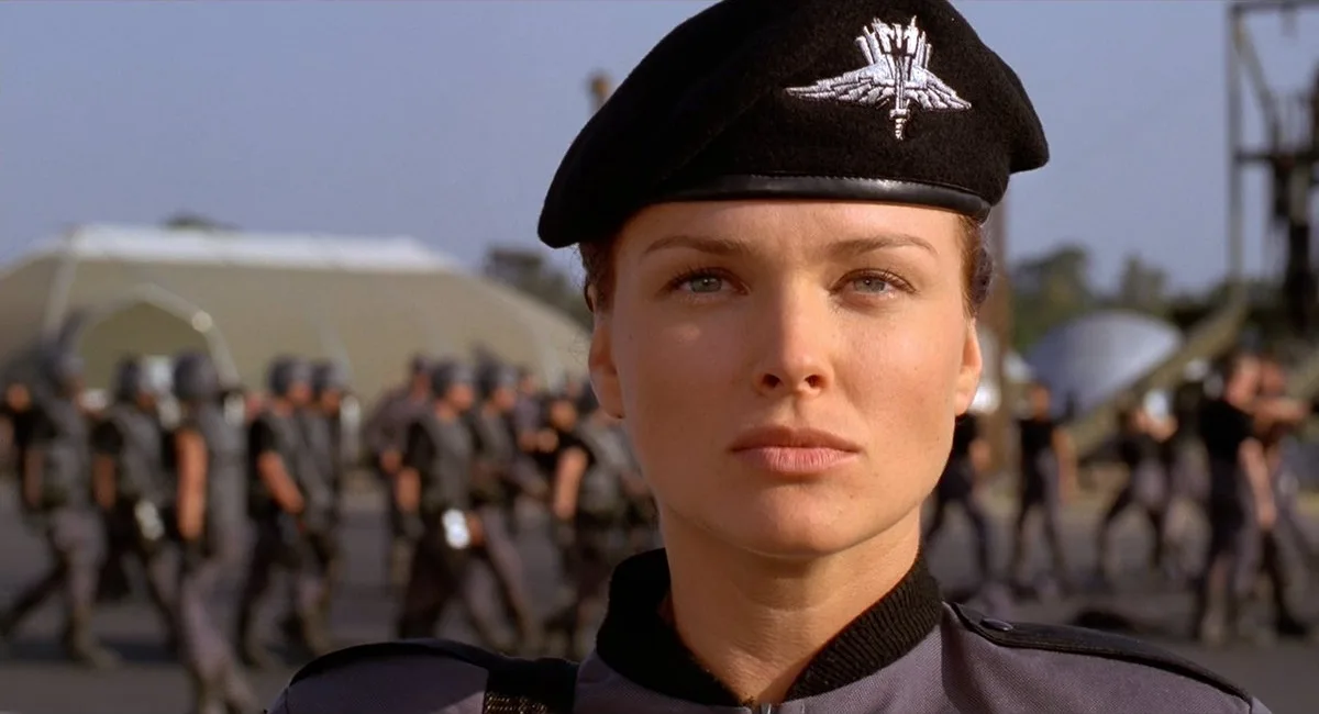 A woman in a black beret with a silver insignia stares intently forward, wearing a dark uniform. Behind her, a blurred group…