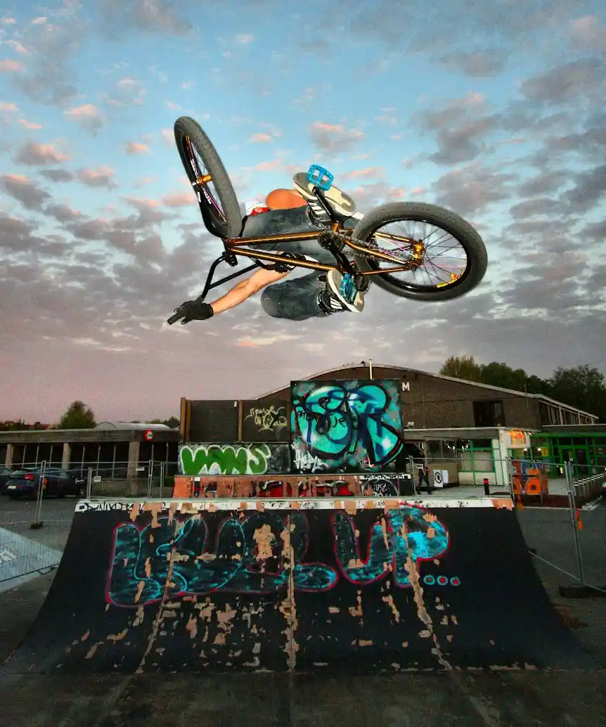 A person is mid-air on a BMX bike performing an inverted trick above a graffiti-covered ramp at dusk. The sky is cloudy with…