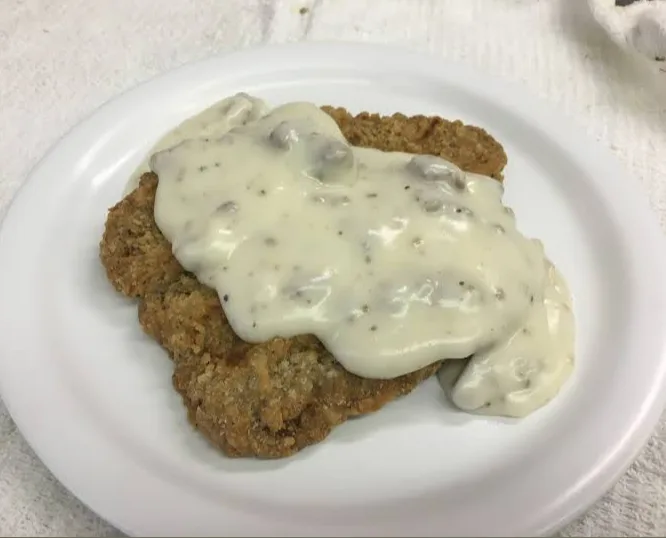 A close-up photo shows a breaded, fried cutlet on a white plate, generously covered with creamy white gravy speckled with…