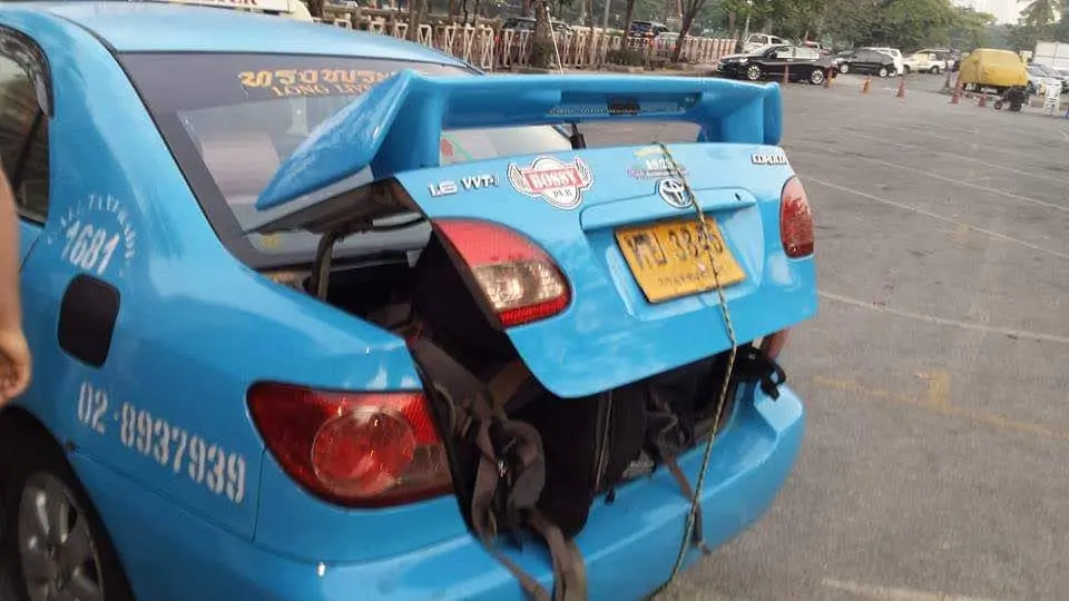 a bright blue taxi with its rear trunk open and a large backpack tied to the back using a rope, parked in a lot. The car has…