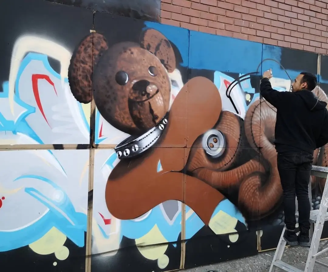 A man in a black hoodie stands on a ladder painting a large mural of a brown teddy bear on a brick wall. The teddy bear has…