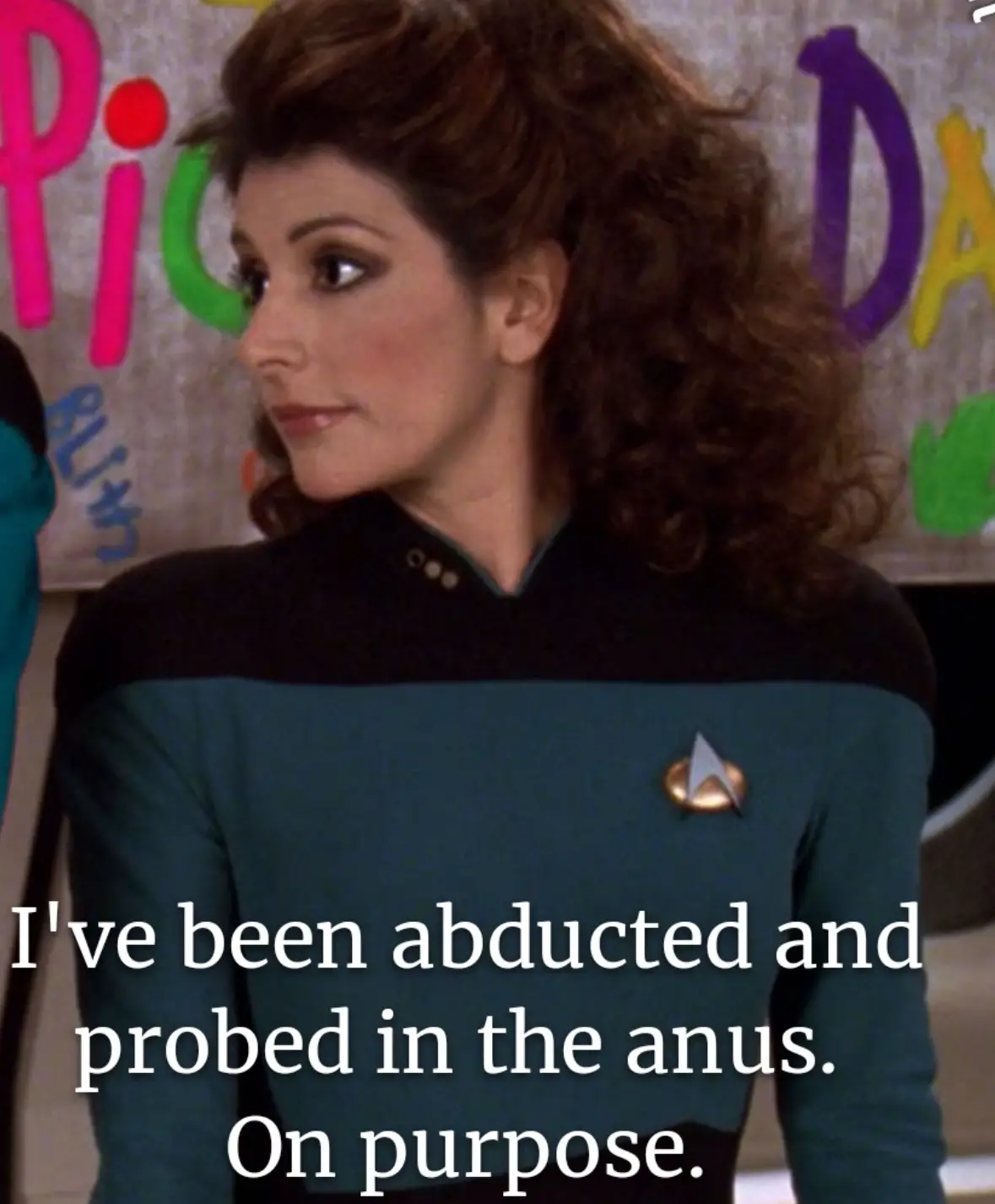A woman in a Star Trek uniform with a calm, slightly amused expression looks off to the side. Behind her, a colorful…
