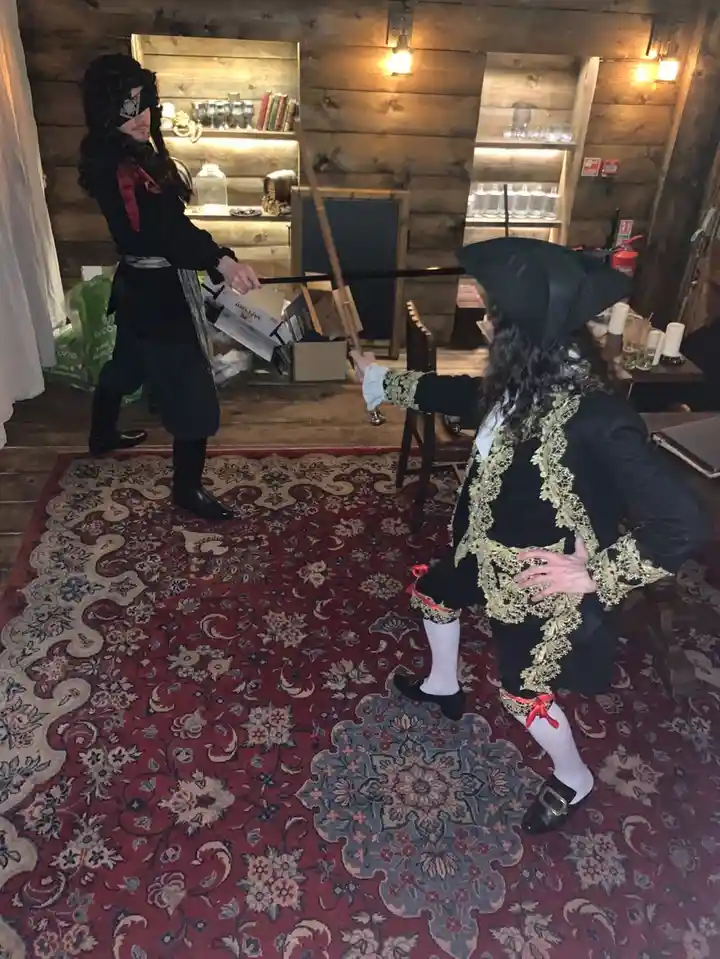 Two people dressed as pirates stand facing each other in a wooden room with shelves and a patterned rug; one holds a sword…