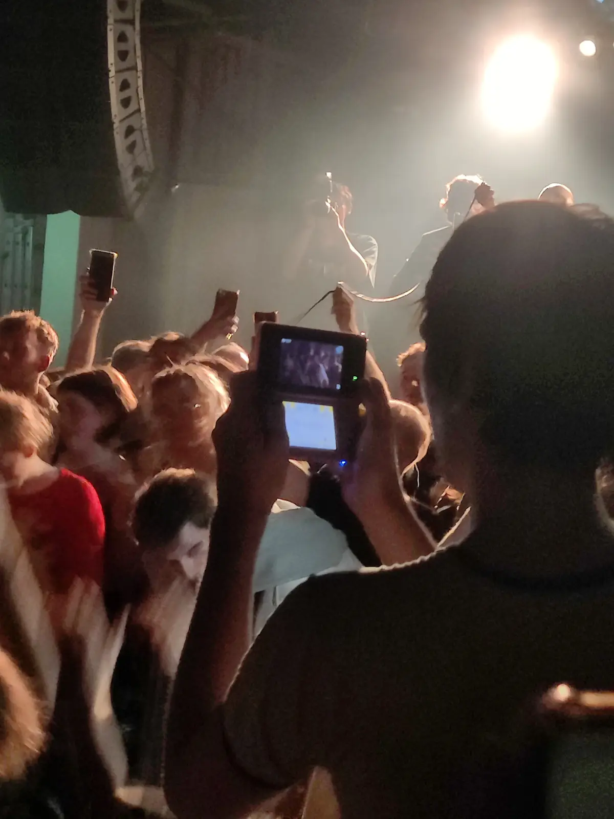 A person at a concert holds up a Nintendo DS, its screen lit up, while the crowd around them raises smartphones to record…