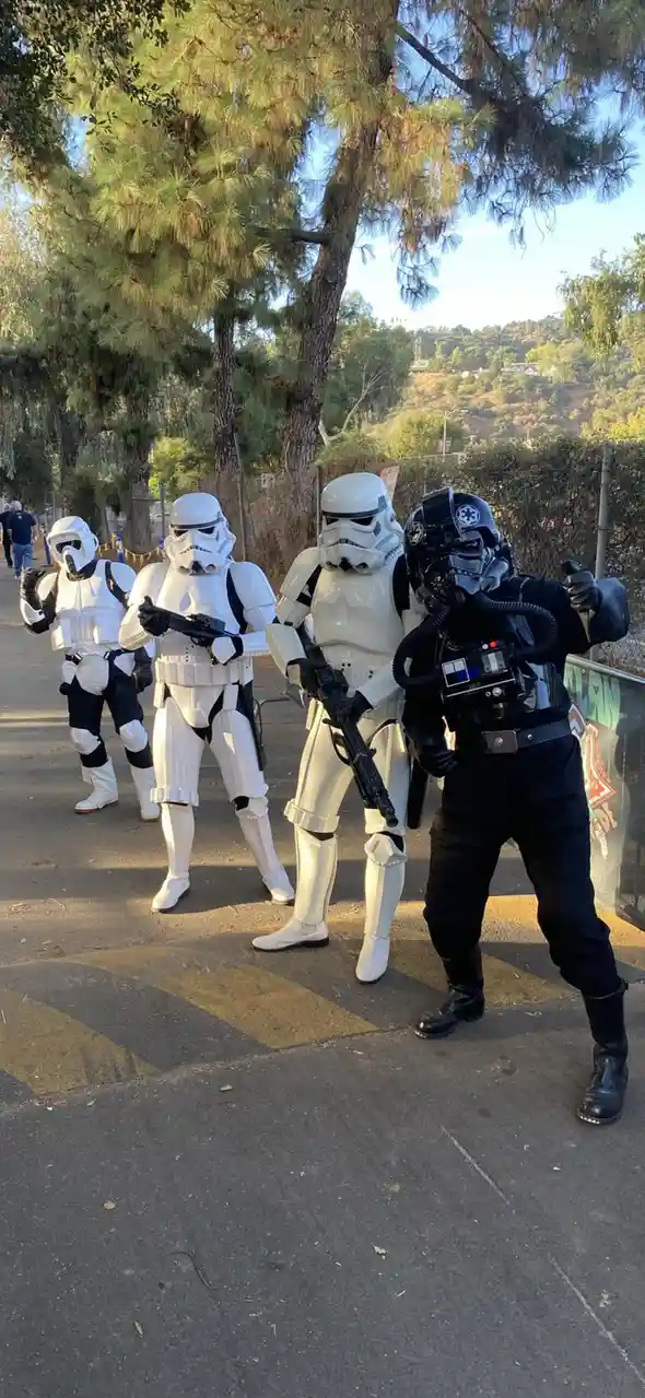 Four people in Star Wars costumes stand outdoors on a paved path under trees. Three are in standard white Stormtrooper armor…