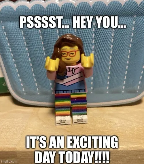 A lego minifigure with brown hair, red glasses, and a rainbow-striped outfit stands on a wooden surface in front of a blue…