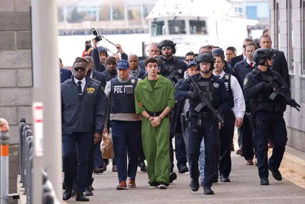 A young man in a green jumpsuit, handcuffed at the wrists, is being escorted by numerous NYPD and tactical police officers…
