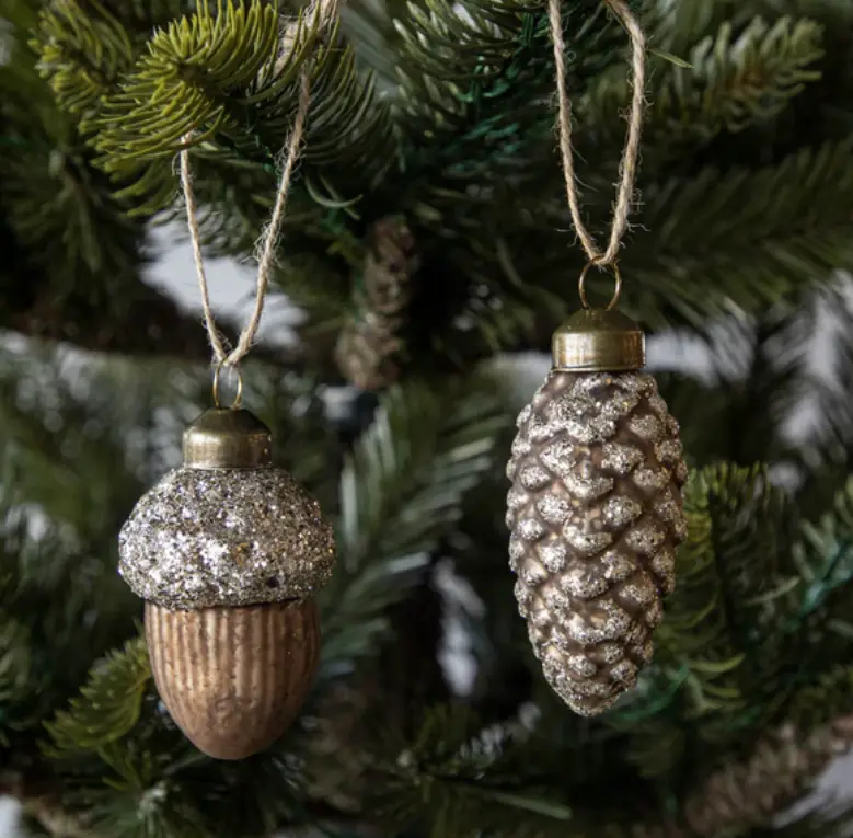 Two glittery Christmas ornaments shaped like an acorn and a pinecone hang from twine on a green pine tree branch. The acorn…