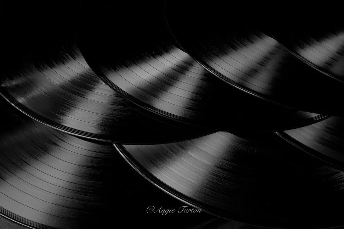 A black-and-white photograph shows several vinyl records stacked and overlapping, their grooves catching light in a moody…