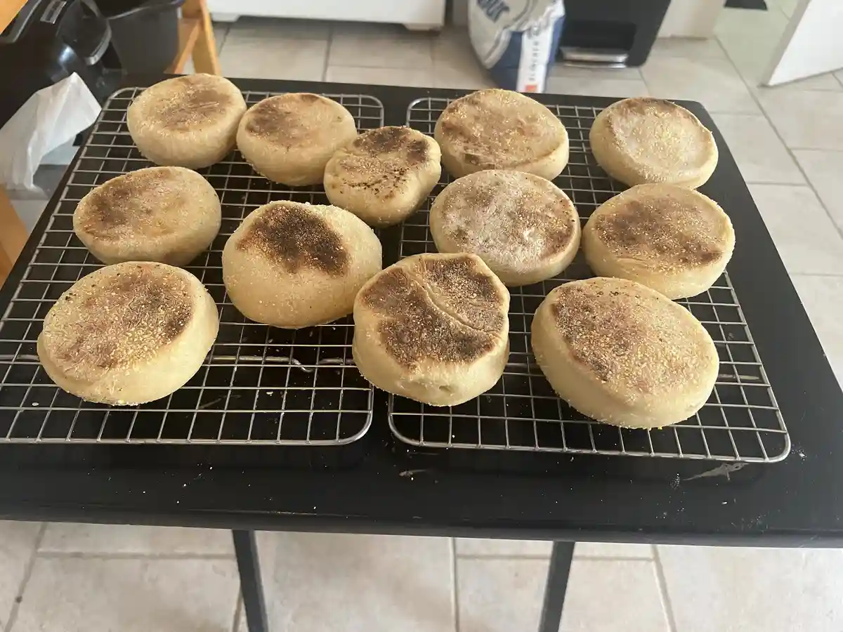 A photo shows a dozen freshly baked English muffins cooling on a wire rack atop a black table in a kitchen. Some muffins…