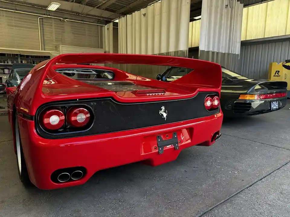 A red Ferrari F50 is shown from the rear in a garage, featuring its iconic large rear wing, circular taillights, and…