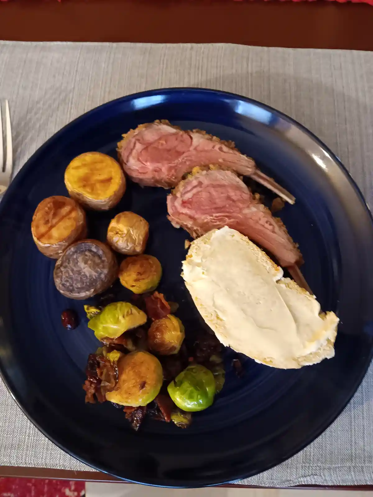 A top-down photo of a dark blue plate holding two cooked lamb chops, roasted potatoes, Brussels sprouts, and a slice of…
