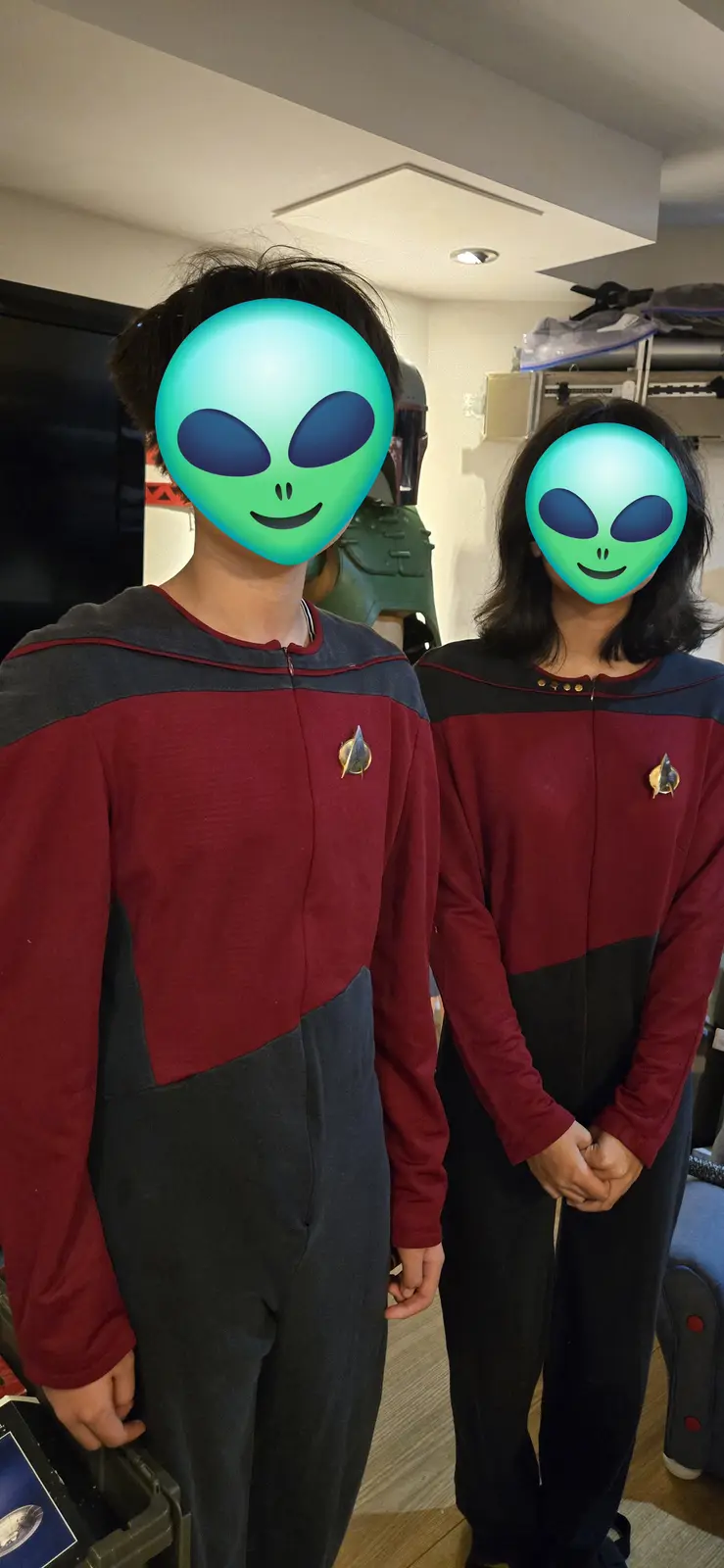 Two people wearing Starfleet uniforms from Star Trek stand side by side indoors, their faces obscured by green alien emoji…