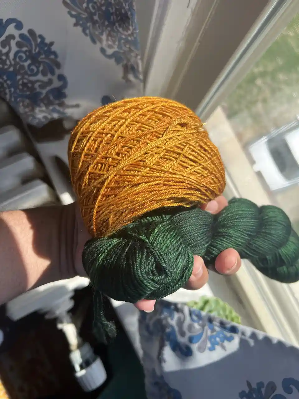 A hand holds two skeins of yarn, one large golden-orange skein shaped like a pumpkin and one smaller dark green skein. The…