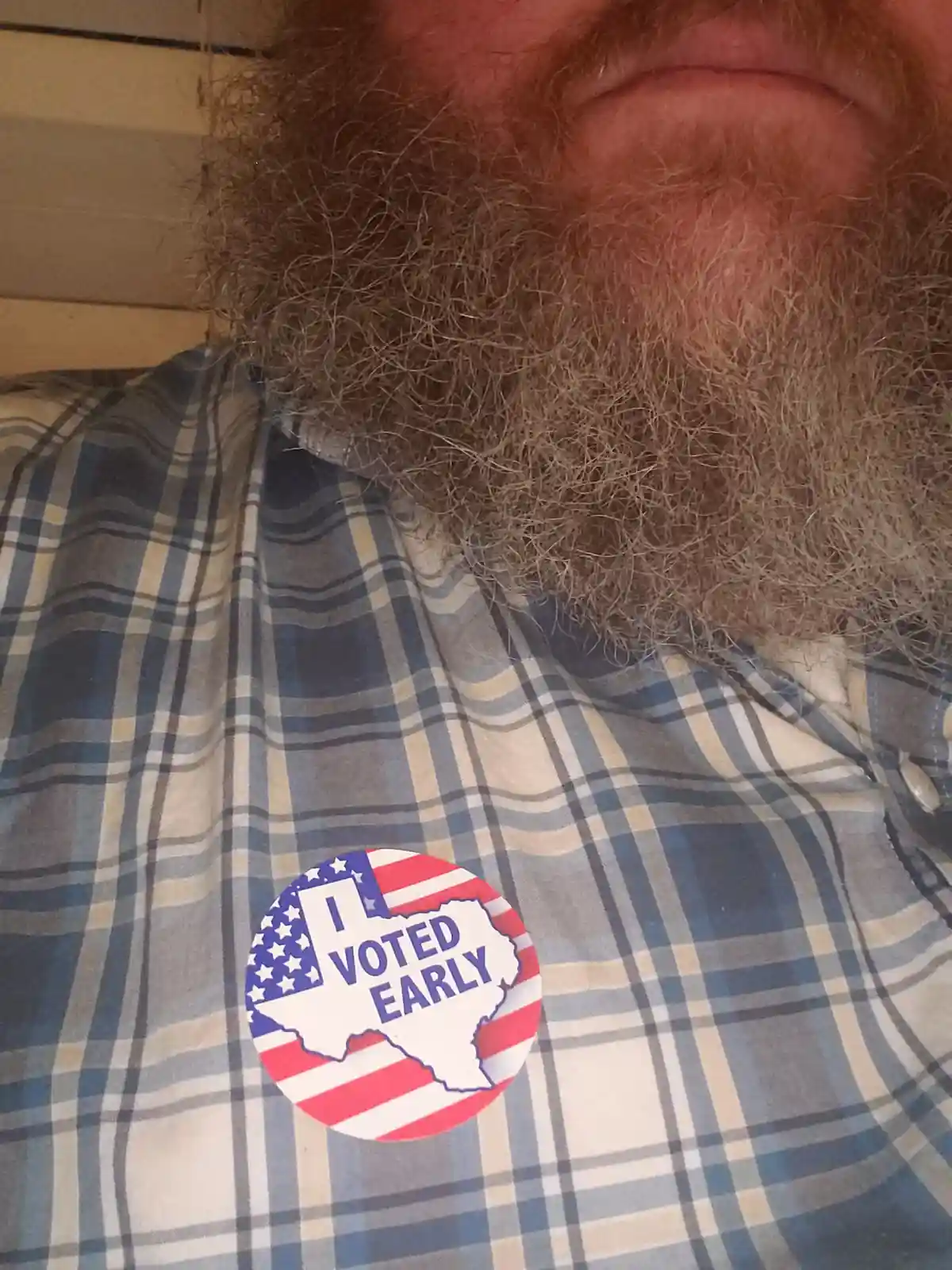 A close-up photo shows a man with a thick, reddish-brown beard wearing a blue, white, and beige plaid shirt. A circular 'I…