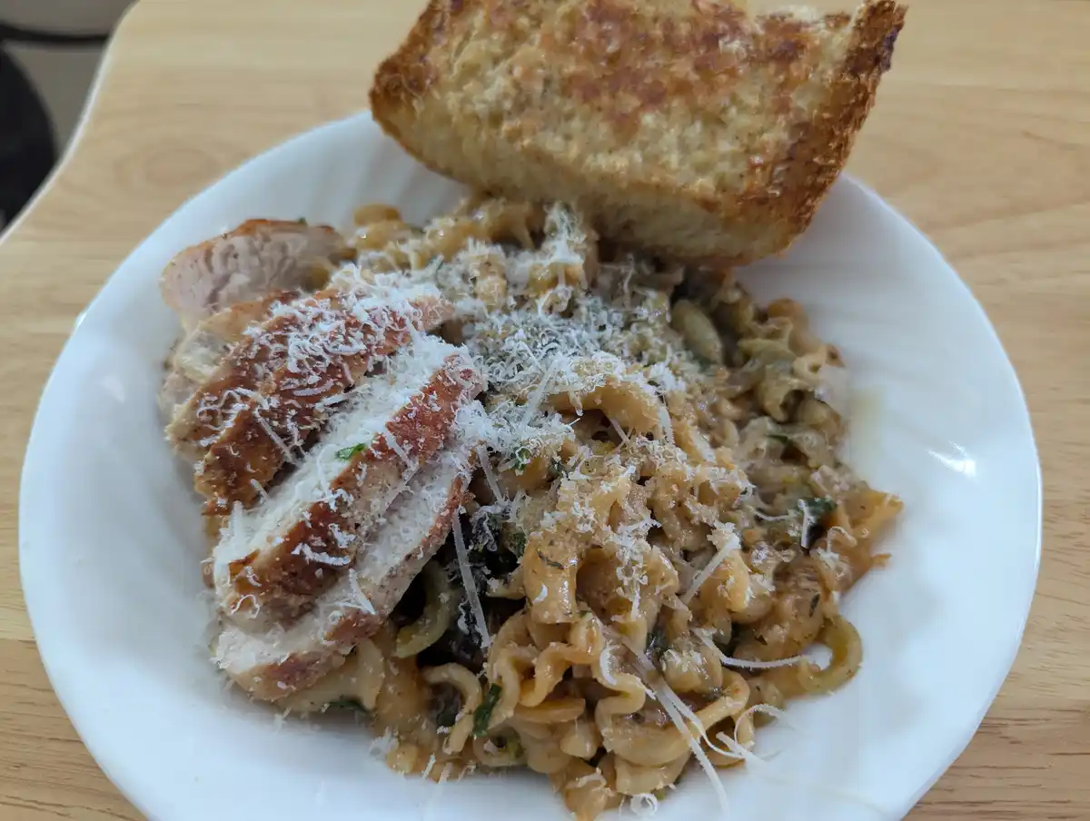 A close-up photo of a white plate holding pasta with sliced chicken and grated cheese, topped with a piece of toasted bread…