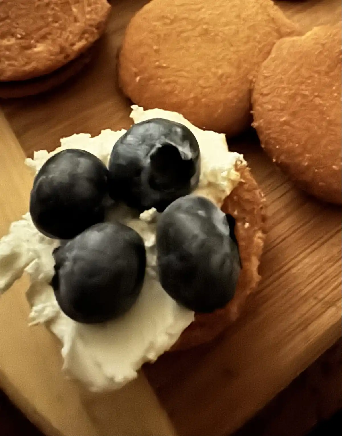 A close-up photo shows four blueberries resting on a dollop of white cream atop a small, round, golden-brown biscuit or…