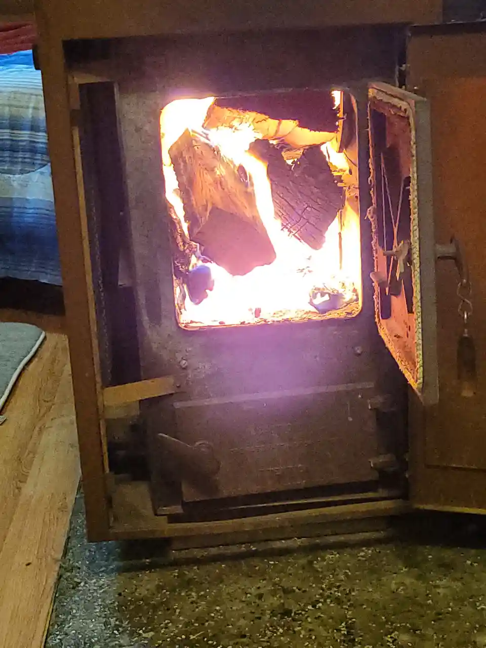 A close-up photo shows the open door of a wood-burning stove with bright, roaring flames and burning logs visible inside…