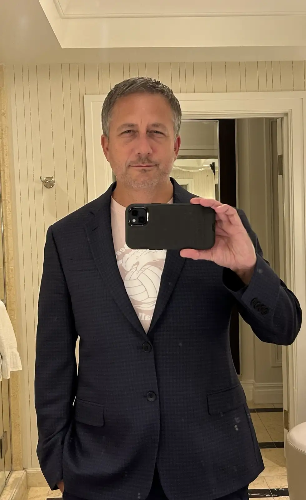 A middle-aged man with short graying hair and a light beard takes a mirror selfie in a well-lit bathroom, wearing a dark…