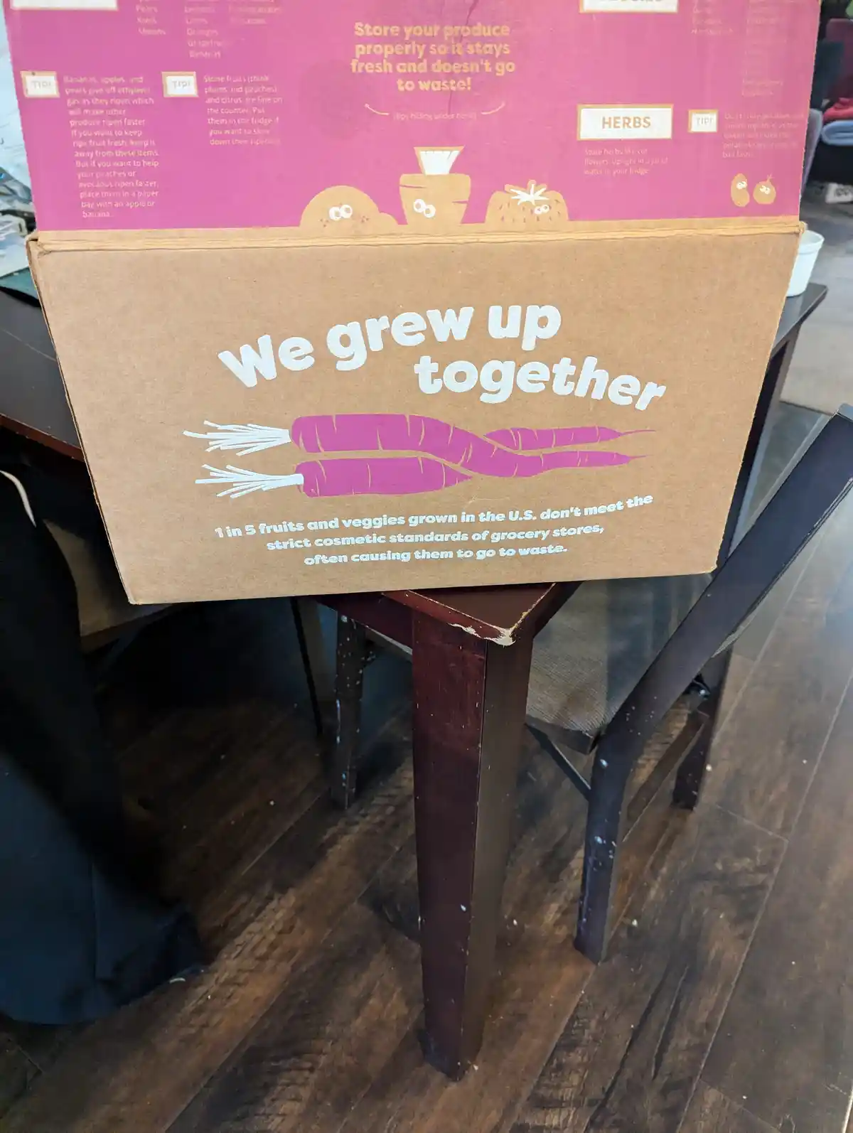 A cardboard box, likely from a produce delivery service, sits on a wooden table. The front of the box features the phrase…