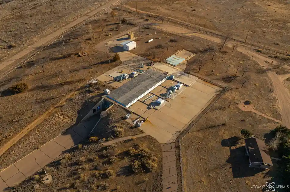 an aerial photograph shows a large, low-profile concrete building with a sloped entrance, surrounded by barren desert…