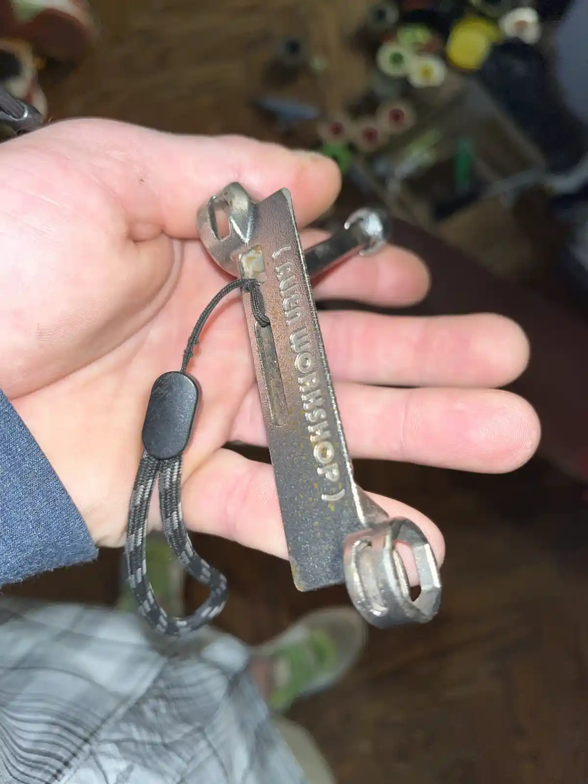 A hand holds a small, worn, multi-tool wrench with 'TURN WORKSHOP)' engraved vertically on its body. The tool has a black…