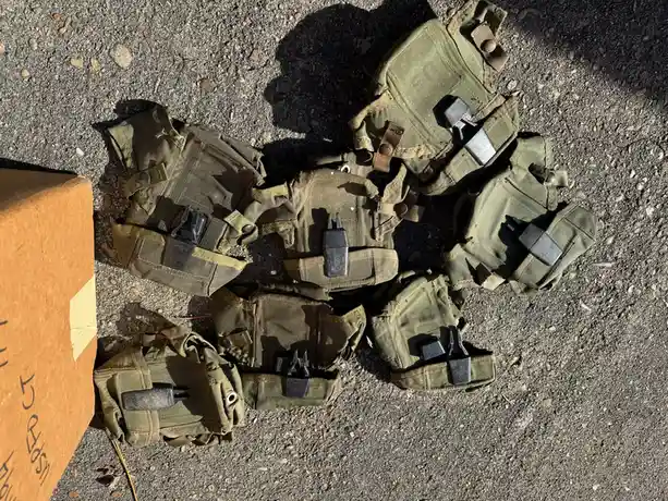 Several olive-drab military-style pouches with black plastic clips are scattered on gravel ground next to a cardboard box…