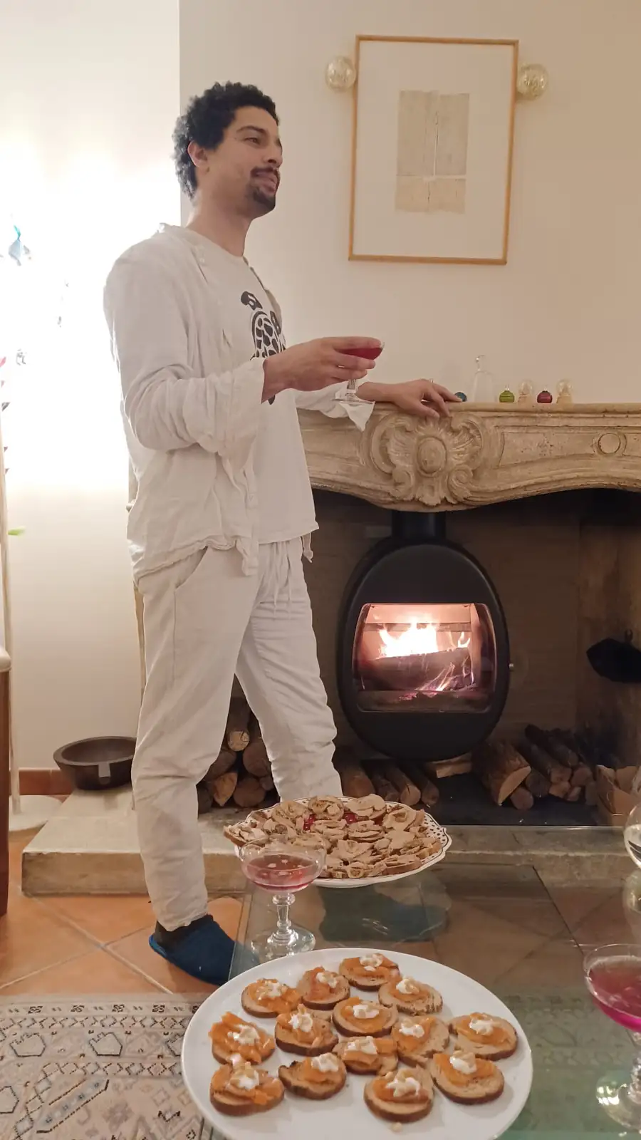 A man in white loungewear stands beside a lit fireplace, holding a glass of red drink, with plates of appetizers on a glass…