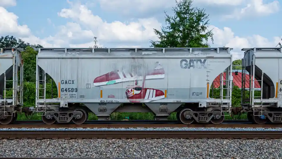 A weathered GATX freight train car with graffiti art of red and white adidas sneakers on its side, parked on railroad tracks…