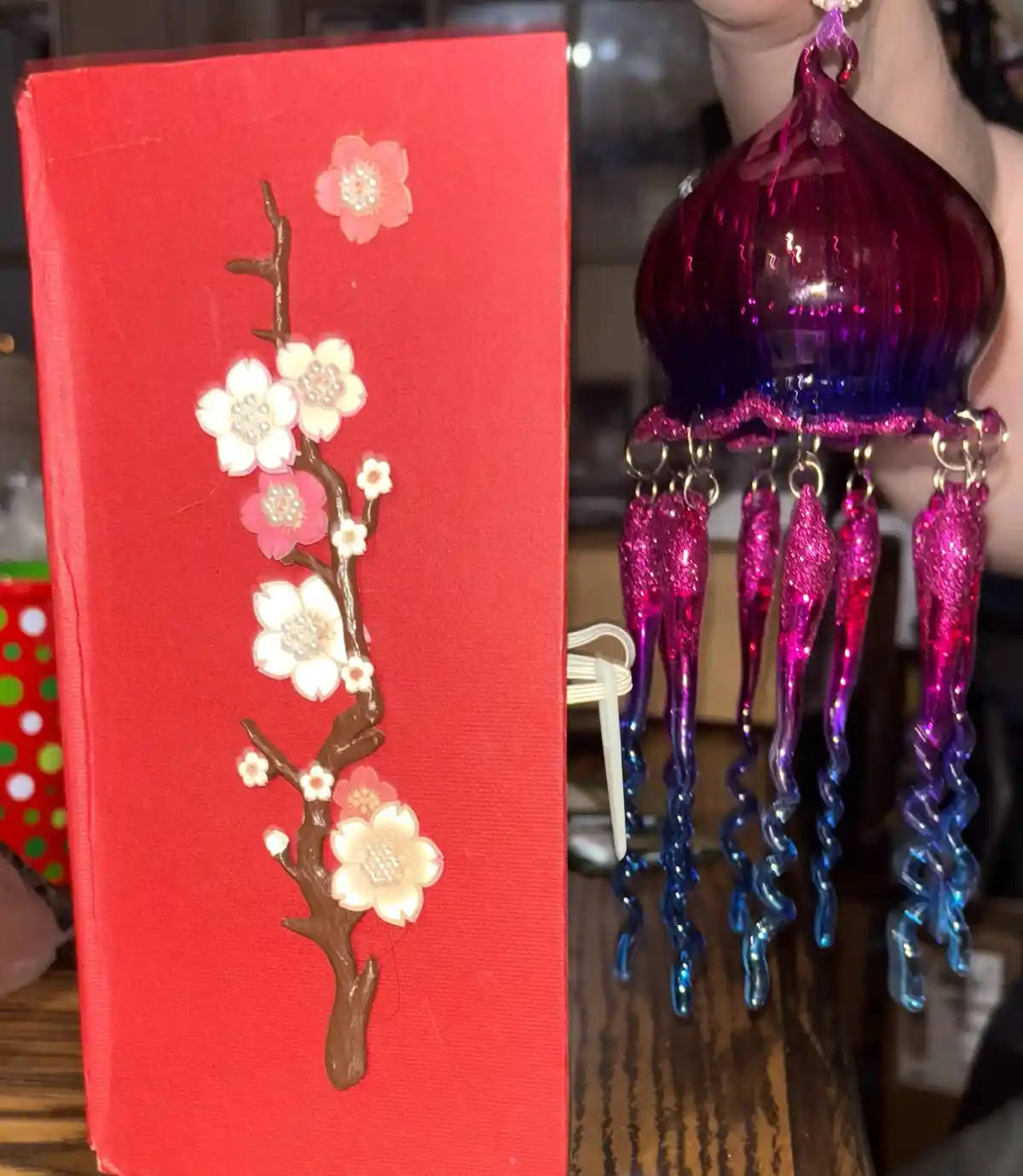A red envelope decorated with a brown cherry blossom branch and pink/white flowers sits beside a shiny purple and blue…