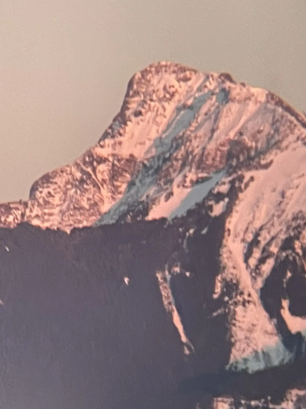 A close-up photo of a snow-covered mountain peak with pinkish-orange light from sunrise or sunset illuminating its ridges…