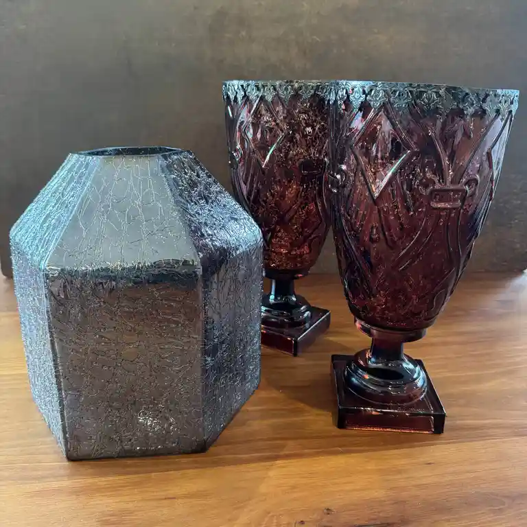 Three decorative glass vases sit on a wooden surface against a dark wall. One is a silver, crackled, hexagonal vase on the…