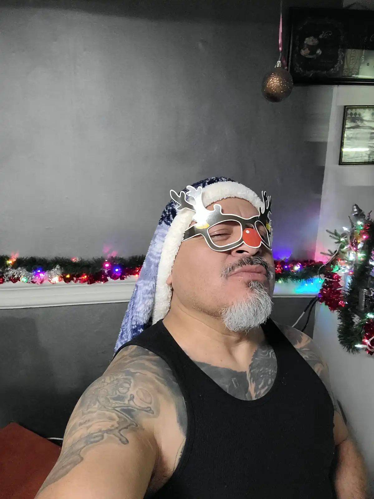 A man with a gray beard and tattoos wears a blue and white Santa hat and reindeer antler glasses with a red nose, looking…