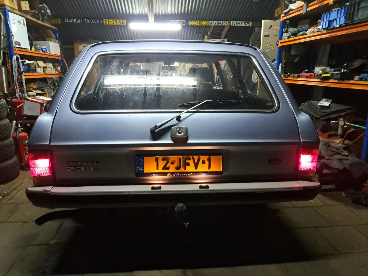 The rear view of a blue Ford Granada 2.3L station wagon parked in a cluttered garage, its brake lights on, with a Dutch…