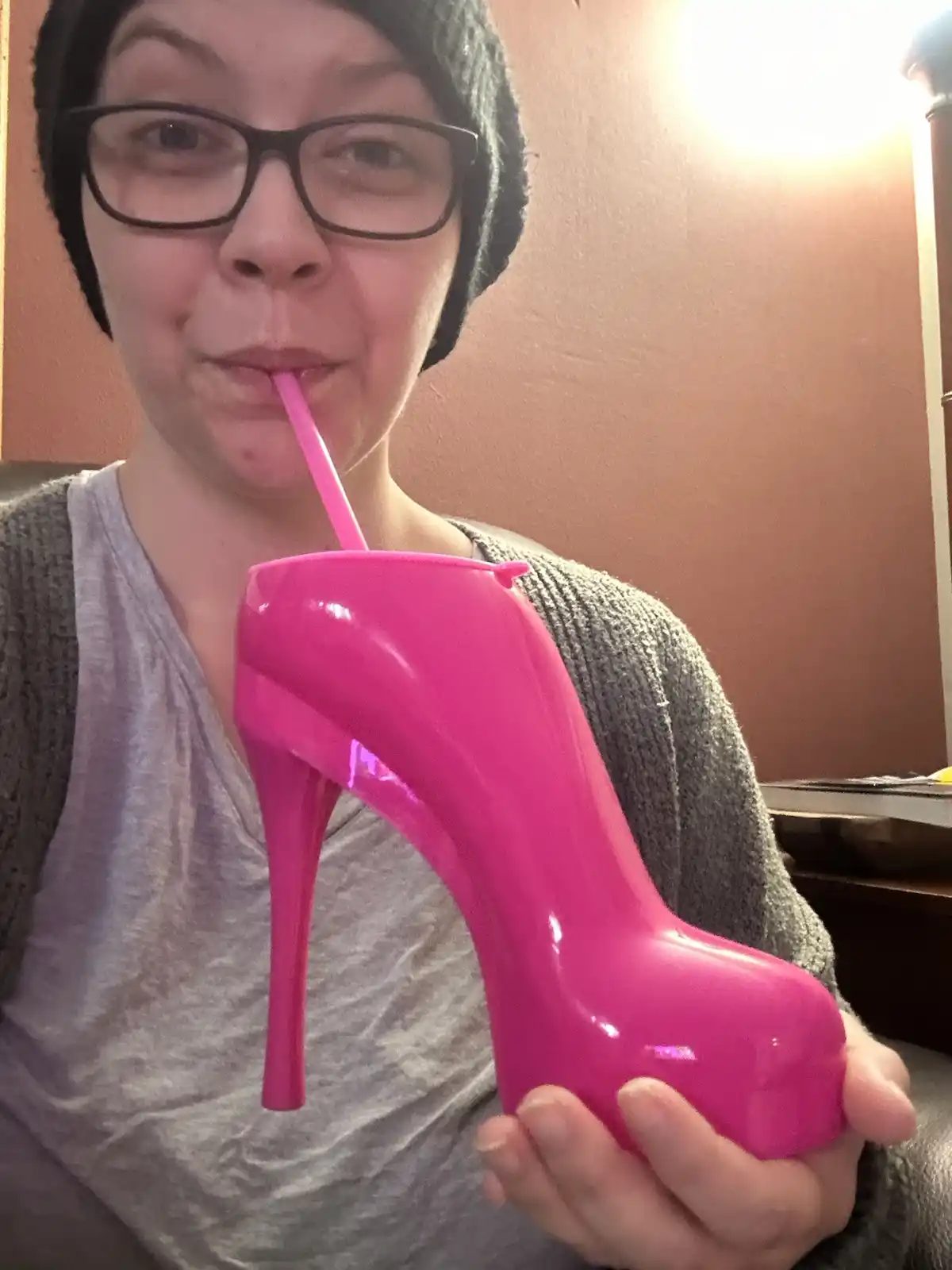 A person wearing glasses and a beanie holds a bright pink plastic high-heeled shoe that has been repurposed as a drink cup…