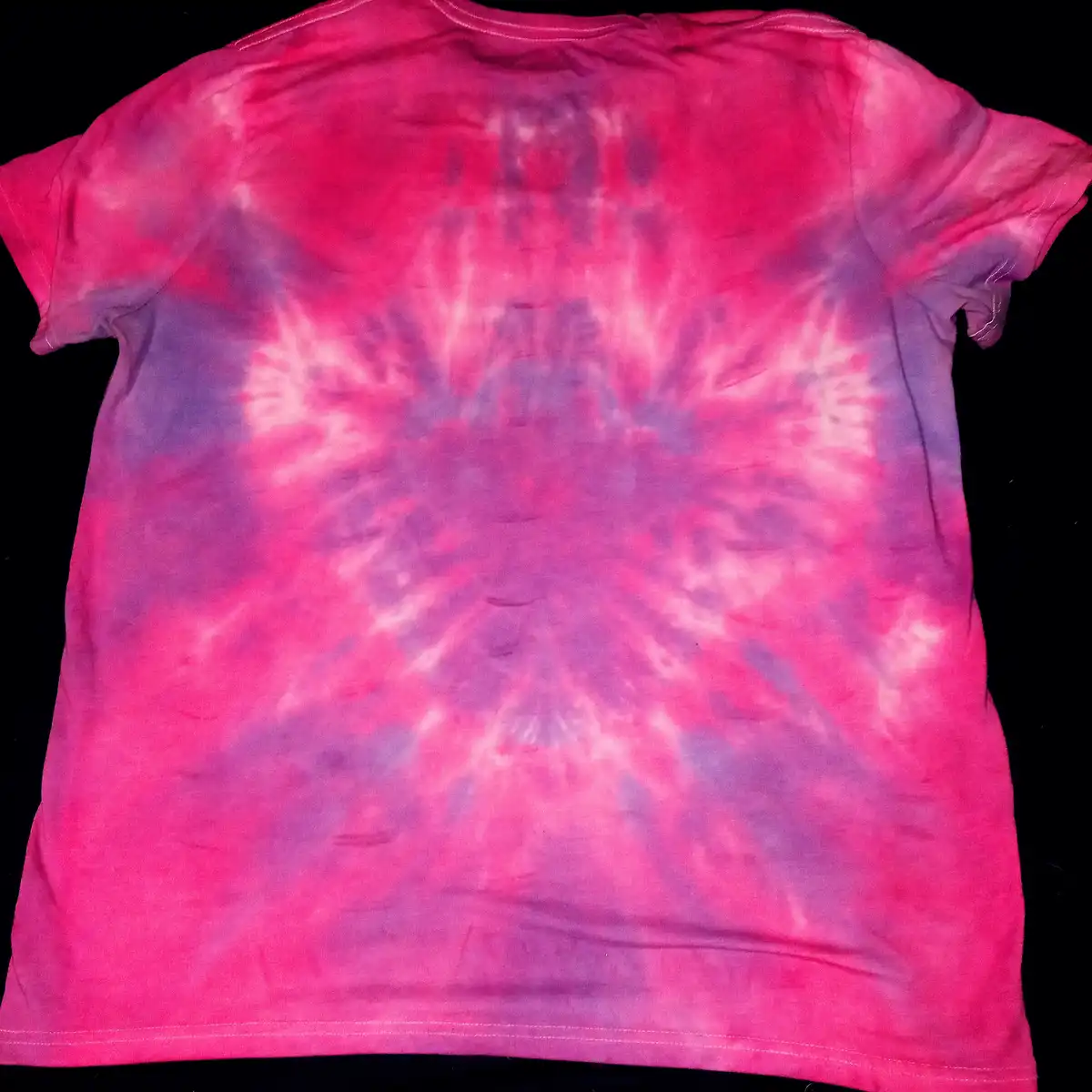 A pink and purple tie-dye t-shirt is laid flat against a black background, showing a radial pattern of dye bleeding outward…