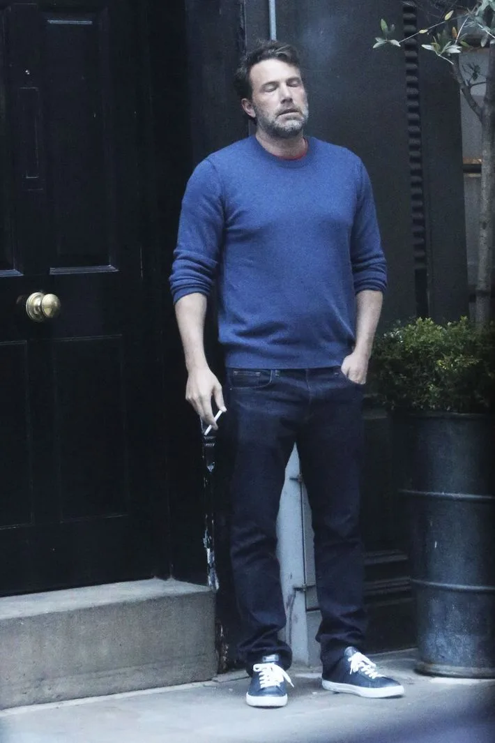 a man in a blue sweater and jeans stands outside a black door, holding a cigarette, with his eyes closed and head tilted…